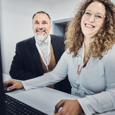 Eine Frau in einem blauen Hemd und ein Mann stehen an einem Schreibtisch vor einem Computer und lachen in die Kamera.