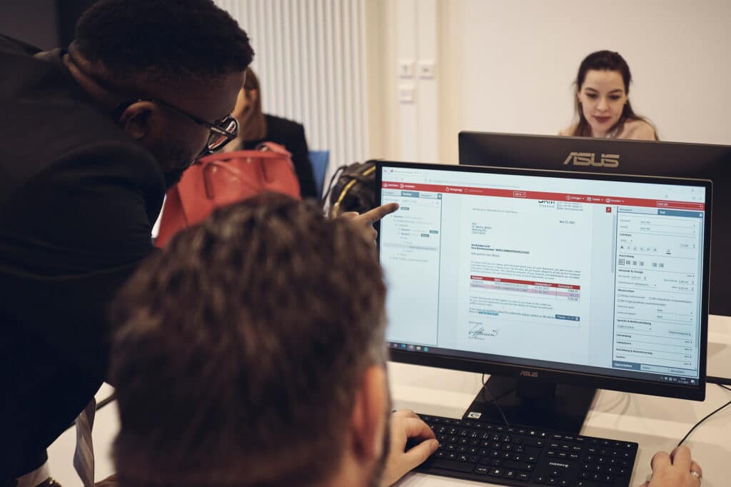 Ein Mann sitzt an einem Computer und verwendet eine Software, während ein anderer Mann in einem Anzug auf den Bildschirm zeigt und etwas erklärt.